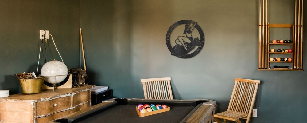 Custom steel sign hung in a man cave room with billiards table and que sticks. Metal sign on back wall in WWII pinup style design with pinup model sitting on large bomb.