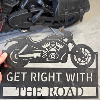 Motorcycle-themed metal sign with 'Get Right with the Road' text held by a person, with a motorcycle in the background.